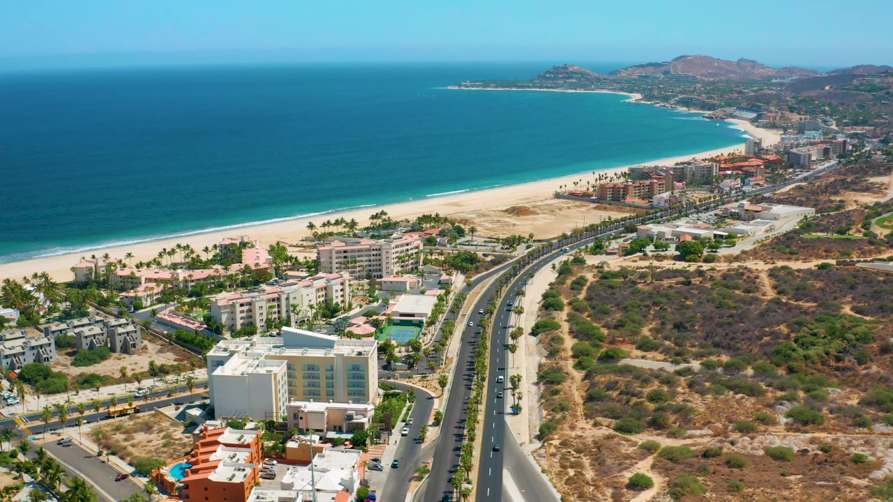 Aerial Los Cabos coastline view with turquoise water at left, a sandy beach curving toward distant hills, hotel buildings near the shore, a divided road below, and no visible people.
