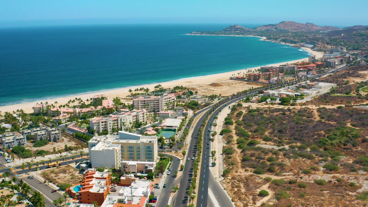 Wide aerial view in Los Cabos, Mexico, showing blue sea, a long sandy beach, coastal roads, low-rise buildings, dry hills, and no visible people.