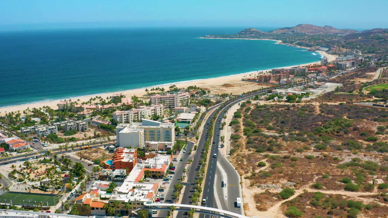 First frame of Los Cabos aerial footage showing turquoise ocean, sandy beach, beachfront buildings, a divided road, dry scrubland, distant hills, and no visible people.