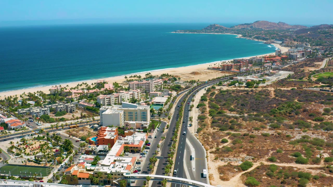 Los Cabos aerial frame with the coastline running across the upper left, urban buildings near the beach, a curving road in the center, scrubland at right, and no visible people.