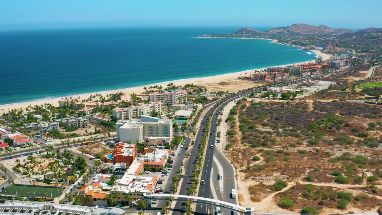 Los Cabos aerial view showing a beachfront neighborhood, pale sand, blue-green ocean, the divided road dropping through the frame, dry open land, and no visible people.