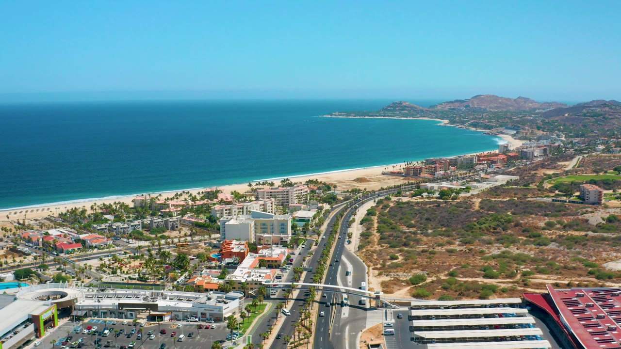 Opening aerial frame over Los Cabos, Mexico, showing blue ocean, a long sandy beach, roadside buildings, dry hills, and no visible people.