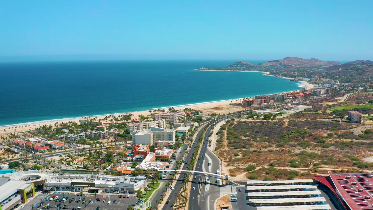 Los Cabos aerial view one second in, with the shoreline curving past low buildings, a divided road, parking lots, dry terrain, and no visible people.