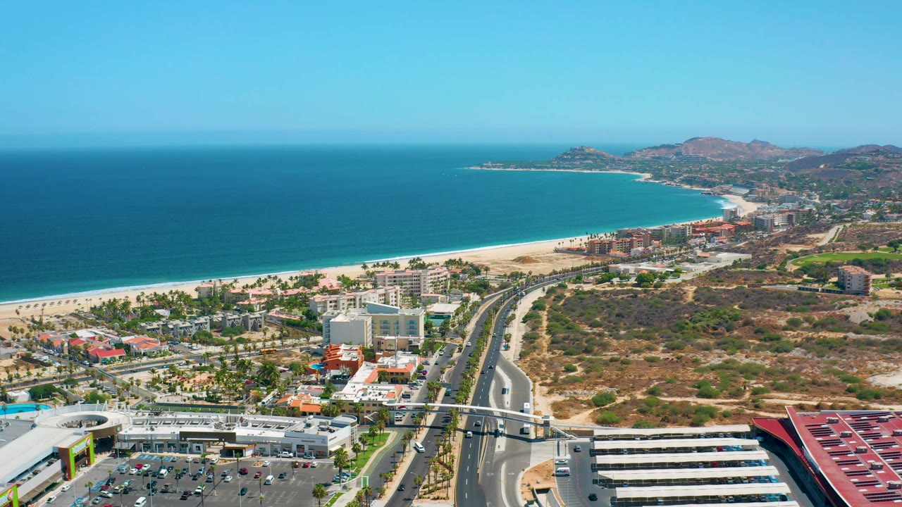 A high daytime frame over Los Cabos, Mexico, with turquoise water to the left, beach below, town buildings near the shore, hills beyond, and no visible people.