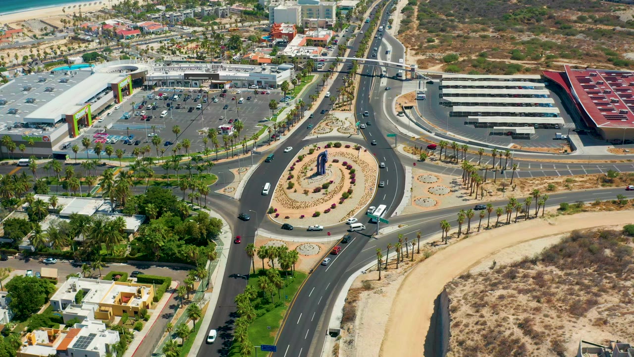 Opening aerial frame over Los Cabos, Mexico, showing a landscaped roundabout, multi-lane roads, parked cars, commercial buildings, private homes, dry lots, and no visible people.