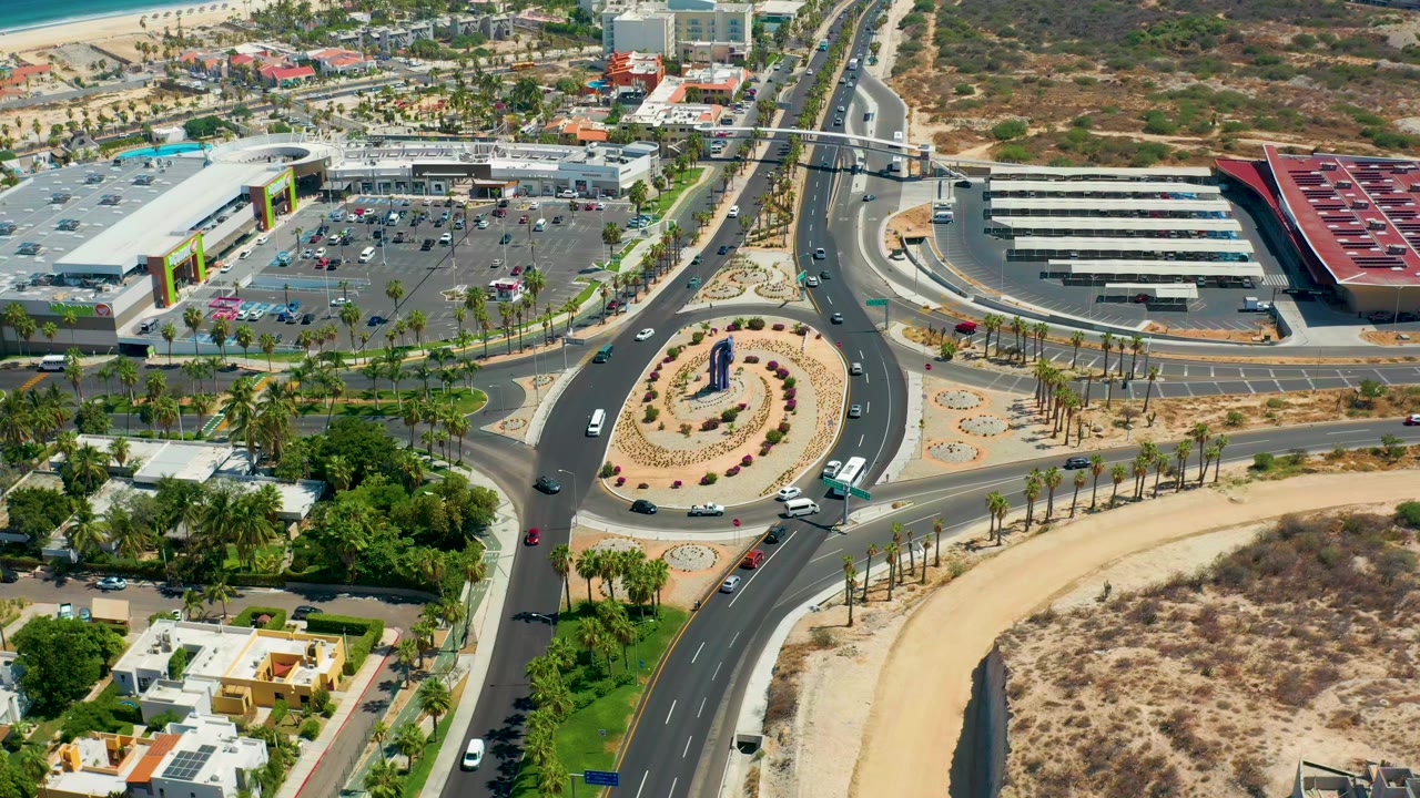 Aerial road junction with cars, commercial roofs, parking areas, palms, and desert lots.