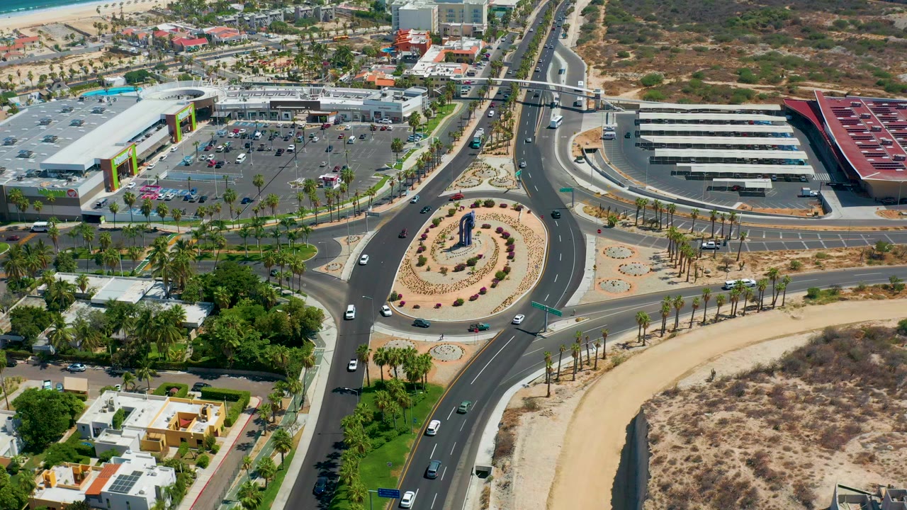 Elevated aerial view in Los Cabos, Mexico, showing a divided highway, circular landscaped roundabout, parking lot, palm trees, modern buildings, and no visible people at the start of the clip.