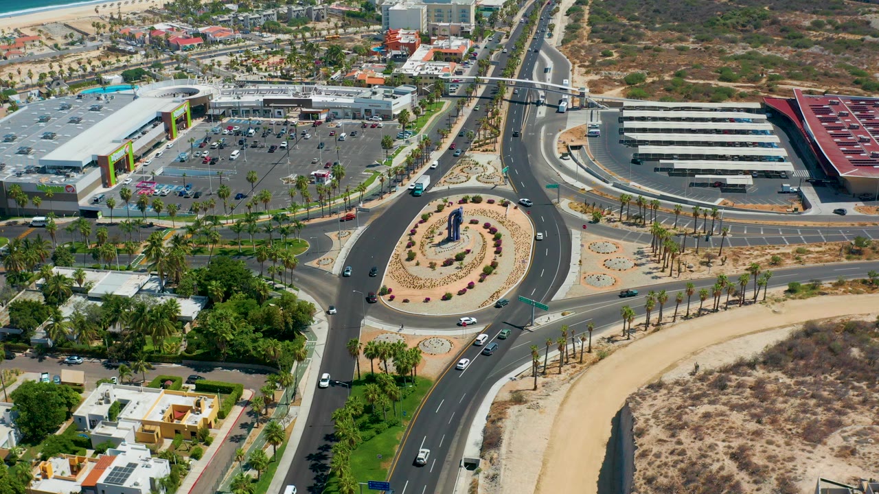 High aerial view in Los Cabos, Mexico showing a landscaped roundabout, curving roads, a parking lot, commercial buildings, desert ground, and no visible people.