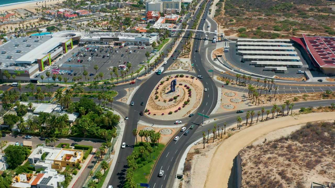 Aerial frame in Los Cabos, Mexico with cars on a curved road around a planted roundabout, large parking areas, roadside palms, dry land, and no visible people.