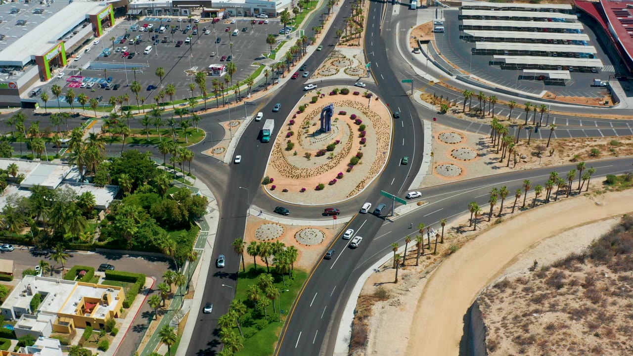 A high aerial view in Los Cabos, Mexico shows a landscaped roundabout, curving roads, cars, parking lots, palms, dry ground, and small pedestrians visible near sidewalks.