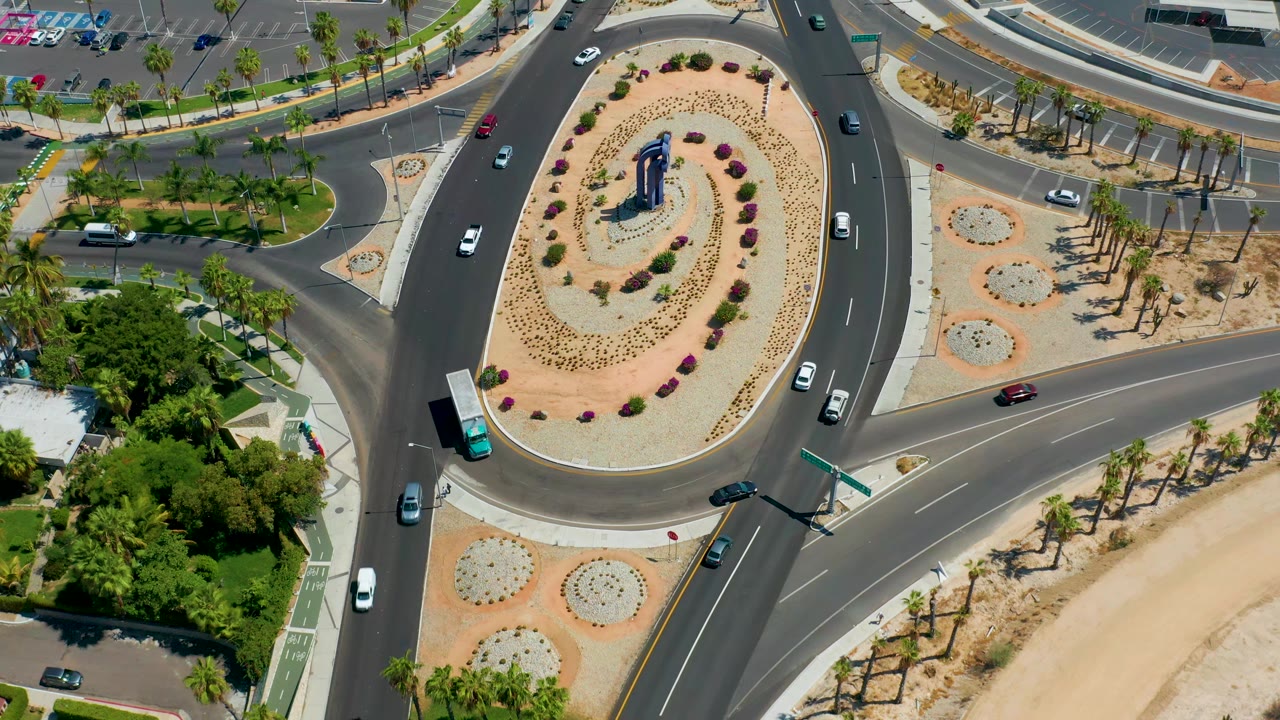 Aerial view over the Los Cabos roundabout as several cars and a turquoise bus occupy different lanes around the oval planted center, with no visible people.