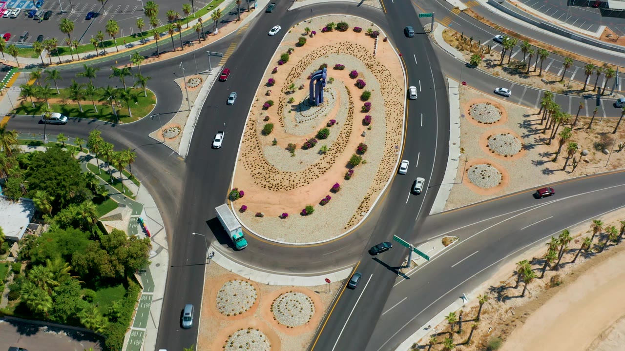 Elevated frame of the Los Cabos traffic circle with cars on the entry and exit roads, a central landscaped island, palms along the road edge, and no visible people.