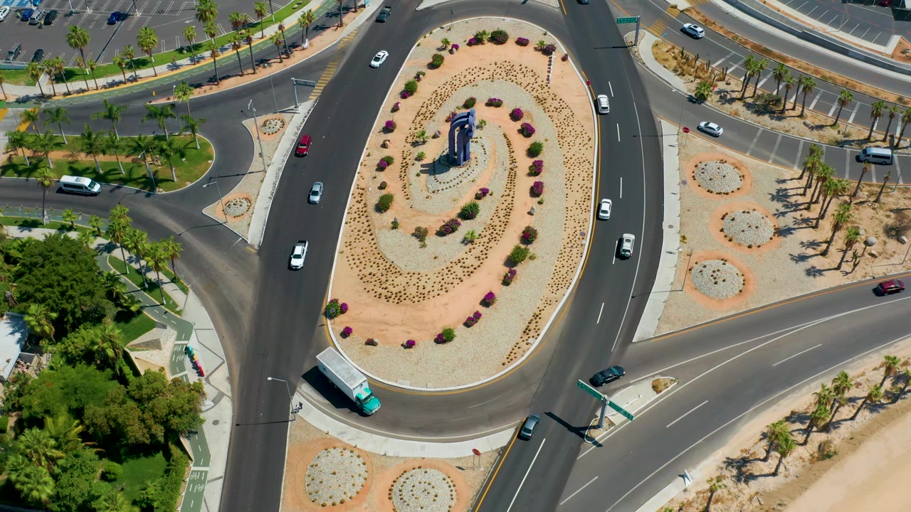 Los Cabos aerial frame with a turquoise bus near the lower edge of the roundabout, cars spaced around the lanes, bright planting beds, and no visible people.