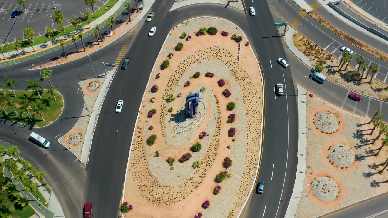 Overhead view in Los Cabos showing curved asphalt lanes, a sandy planted median, several cars, palm-lined sidewalk edges, and no visible people at the start of the clip.