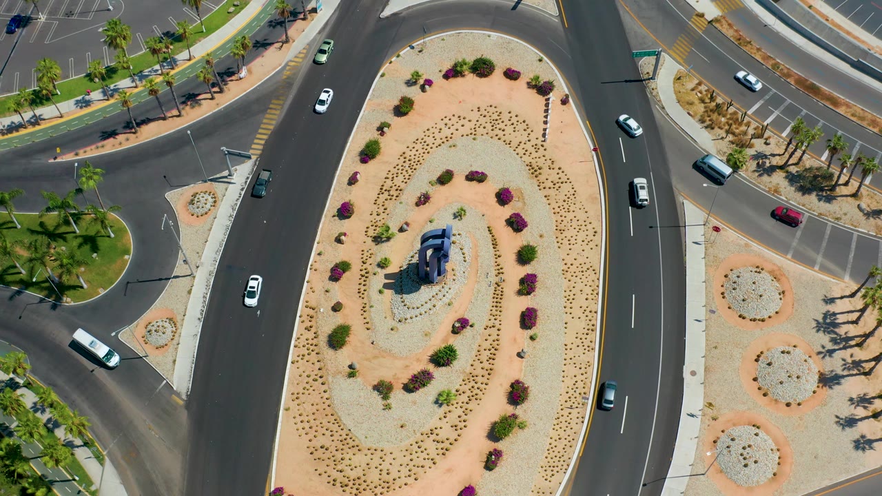 Top-down Los Cabos view with the median shifted through frame, visible cars on both sides of the road, palm shadows near sidewalks, parking spaces, and no visible people.