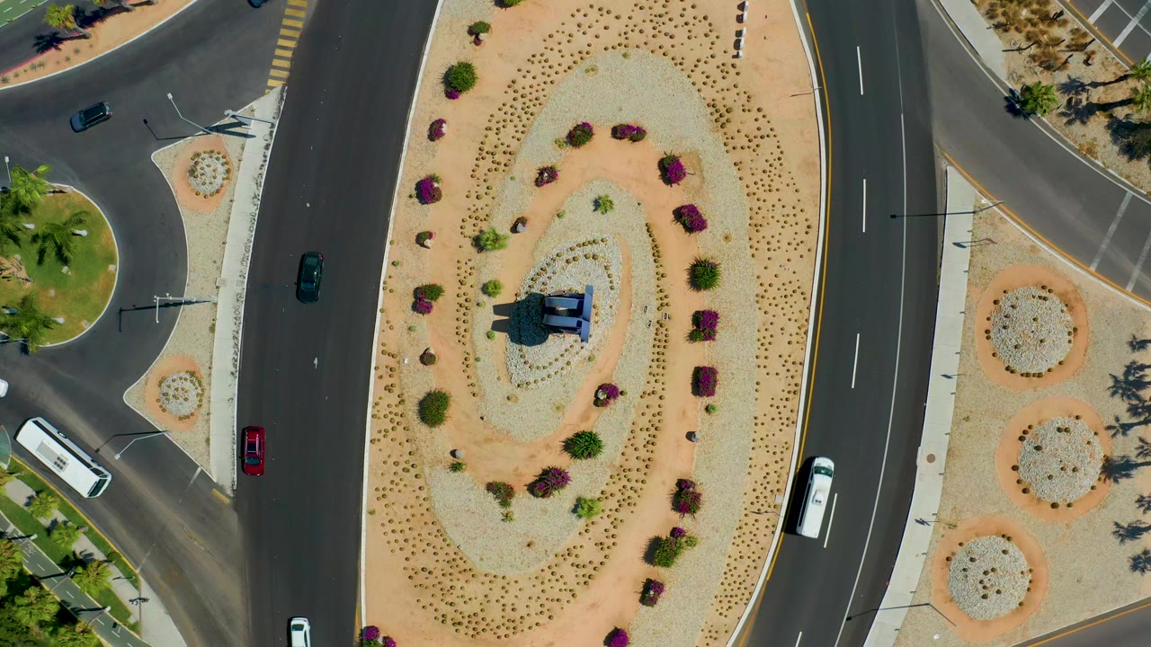High overhead Los Cabos frame showing the oval gravel median, small flowering shrubs, a white bus near the lower right, dark cars on the lanes, sidewalks, palms, and tiny visible people.