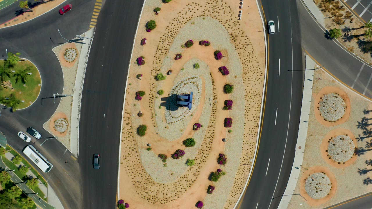 Overhead frame of a Los Cabos, Mexico roundabout with dark roads, a sandy planted oval, a central statue, several vehicles, a bus at lower left, and no visible people.