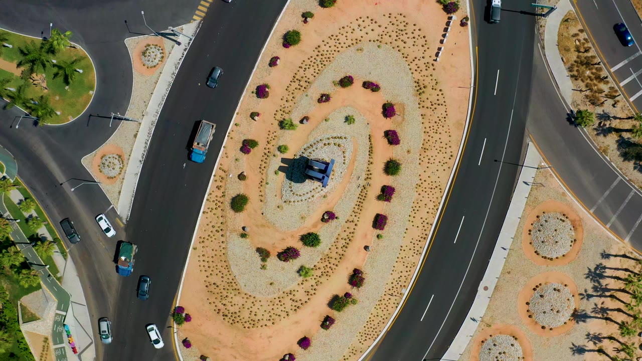 Overhead Los Cabos road frame with vehicles spaced around the roundabout, pale gravel landscaping, curved asphalt lanes, and no visible people.