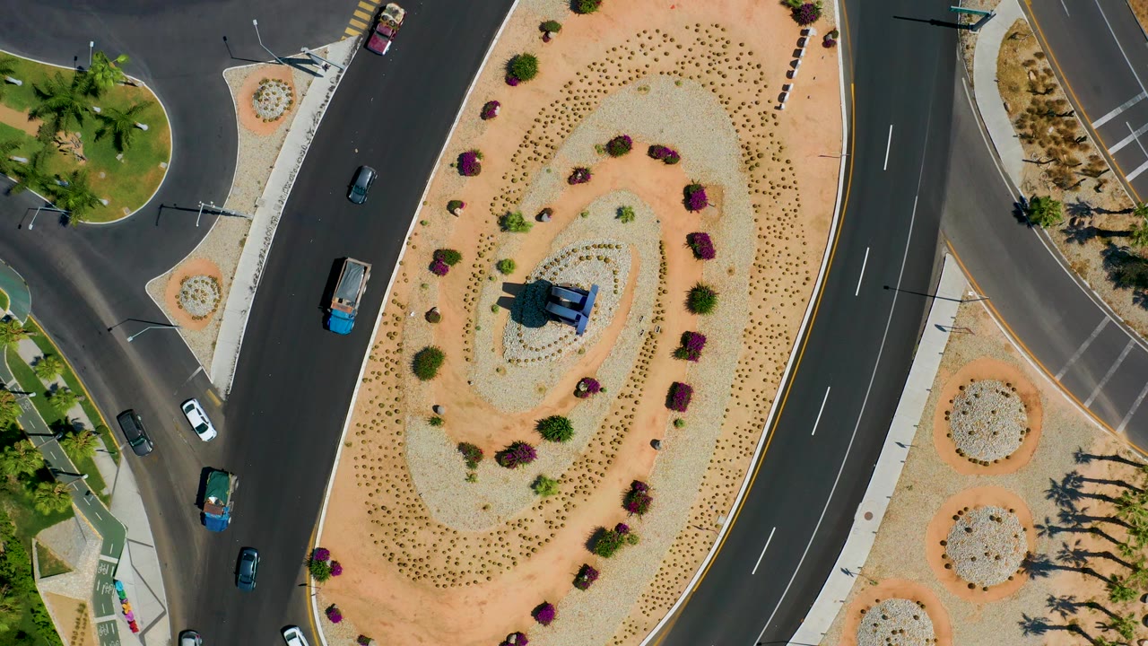 Top-down Los Cabos view of a road junction wrapping a landscaped center island, with several cars, planted shrubs, and no visible people.