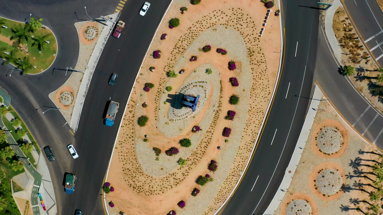 Overhead Los Cabos frame with a white car, a small truck, and darker vehicles moving beside a tan gravel roundabout, with no visible people.