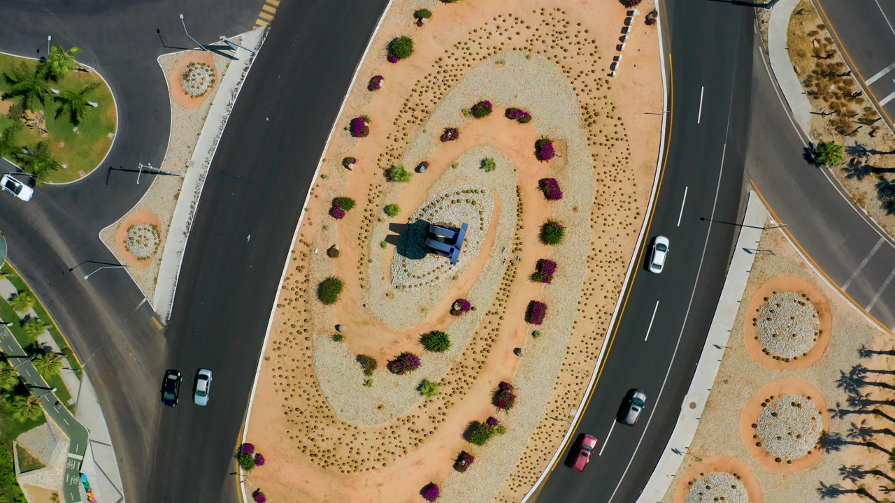 Straight-down aerial frame in Los Cabos, Mexico showing a landscaped oval roundabout between dark asphalt roads, with several cars and no discernible pedestrians.
