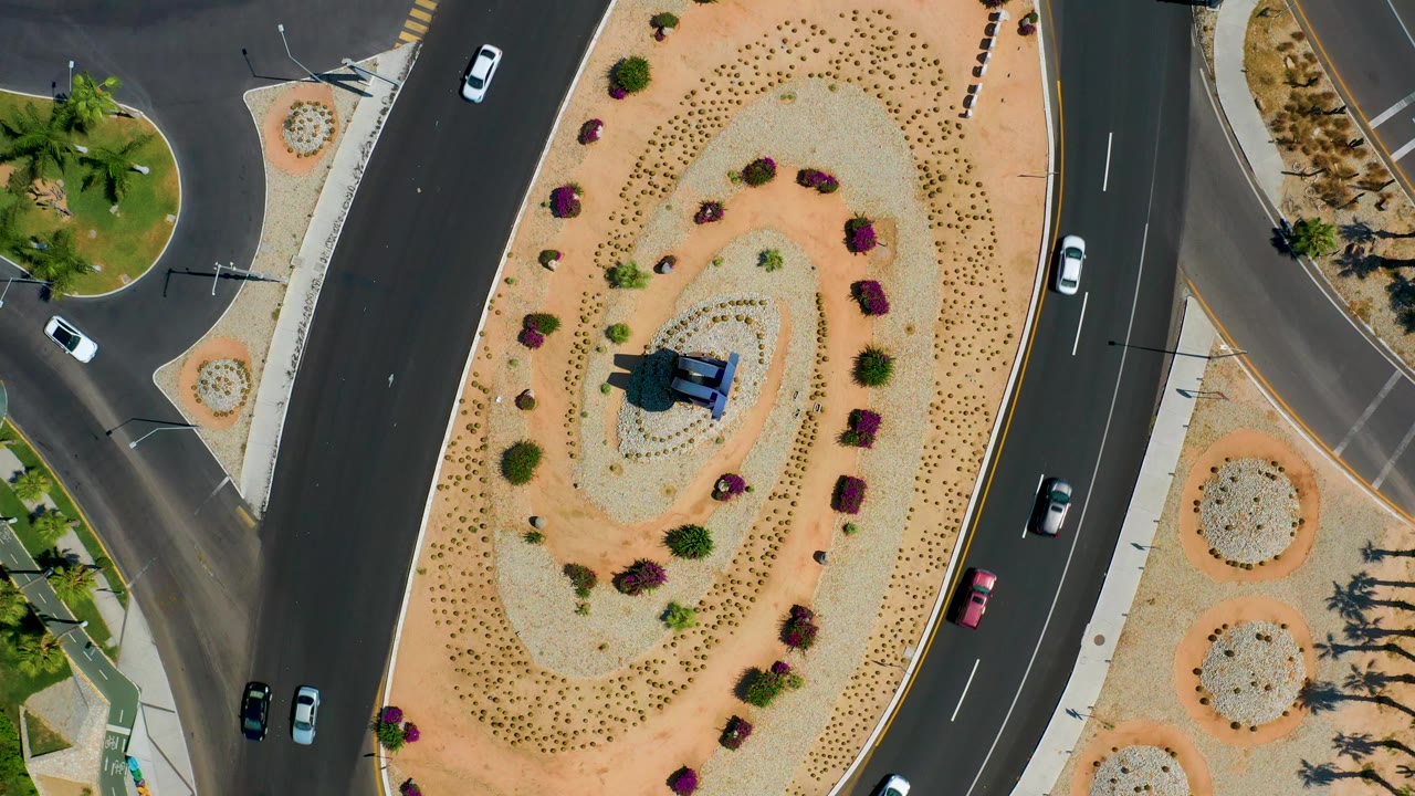 Top-down frame in Los Cabos, Mexico with cars spaced across curved blacktop lanes, desert gravel planting beds, palms, sidewalks, and no discernible pedestrians.