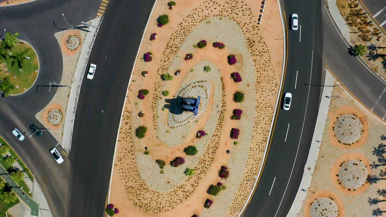 Straight-down aerial frame of a landscaped road island in Los Cabos, Mexico, with curved asphalt lanes, several cars, desert gravel planting, and no visible people.