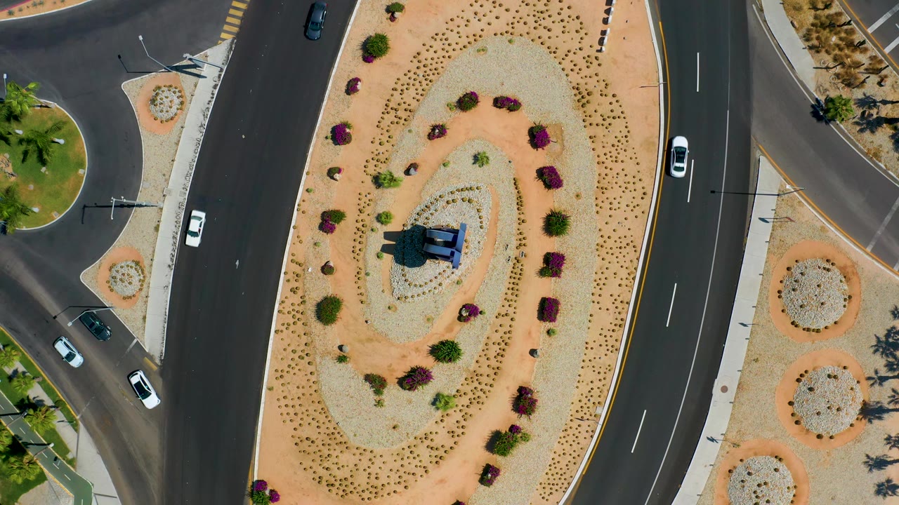 Straight-down aerial view in Los Cabos, Mexico, showing an oval gravel island, patterned shrubs, cars on the left and right roadways, and no visible people.