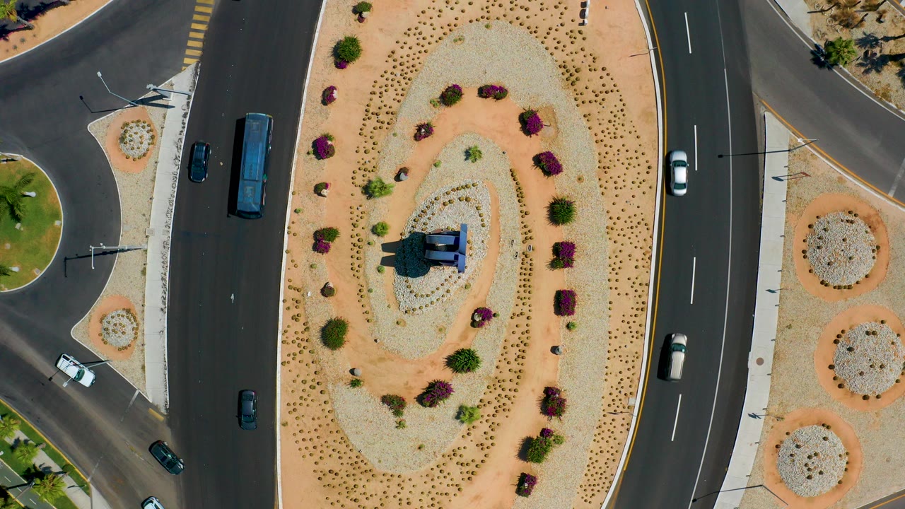 Overhead Los Cabos road view with a curved traffic lane, tan landscaped island, desert shrubs, a central statue, several vehicles, and no visible people.