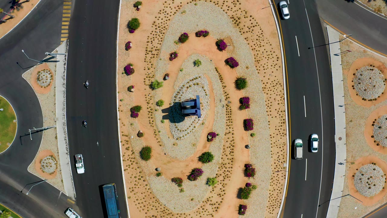 Top-down Los Cabos road frame with the sandy oval planting area centered, dark asphalt lanes on both sides, several cars, scooters, and no visible people.
