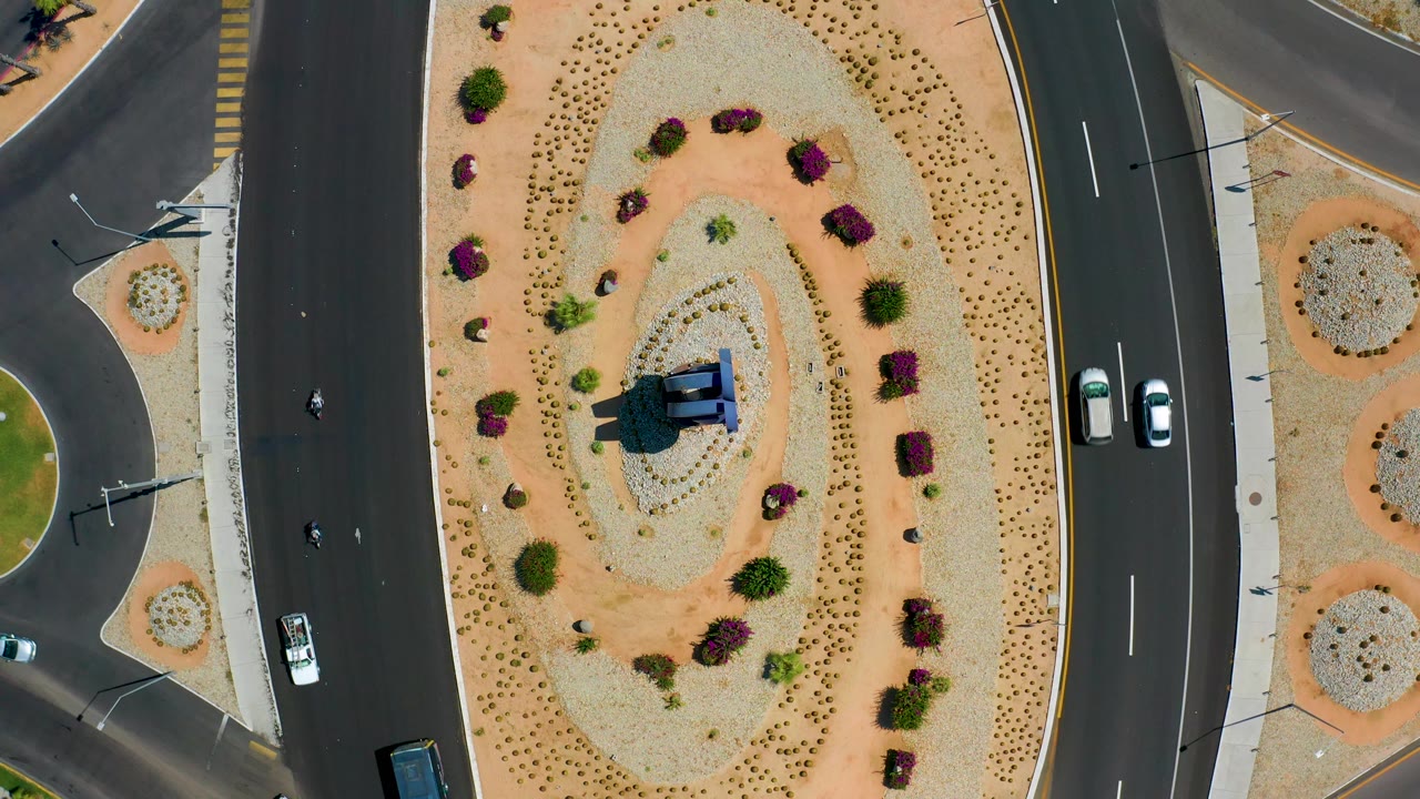Overhead Los Cabos frame showing cars shifting along the right-hand roadway beside a broad sand-and-gravel median with shrubs, rocks, and no visible people.