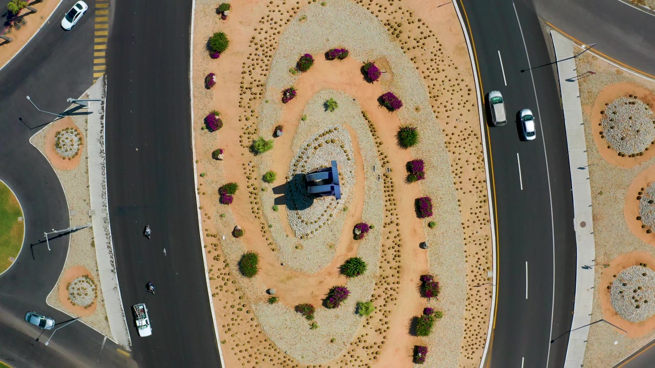 Los Cabos aerial frame with a central sandy landscaped island, purple shrubs, small green plants, dark roads, cars, scooters, and no visible people.