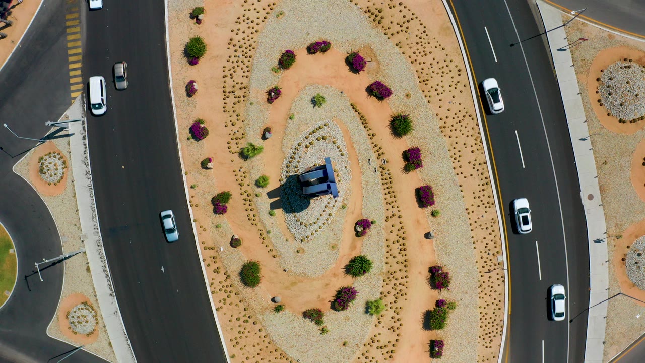 Overhead view in Los Cabos, Mexico of cars traveling on curved roads beside an oval sand-and-gravel median with shrubs and no visible people.