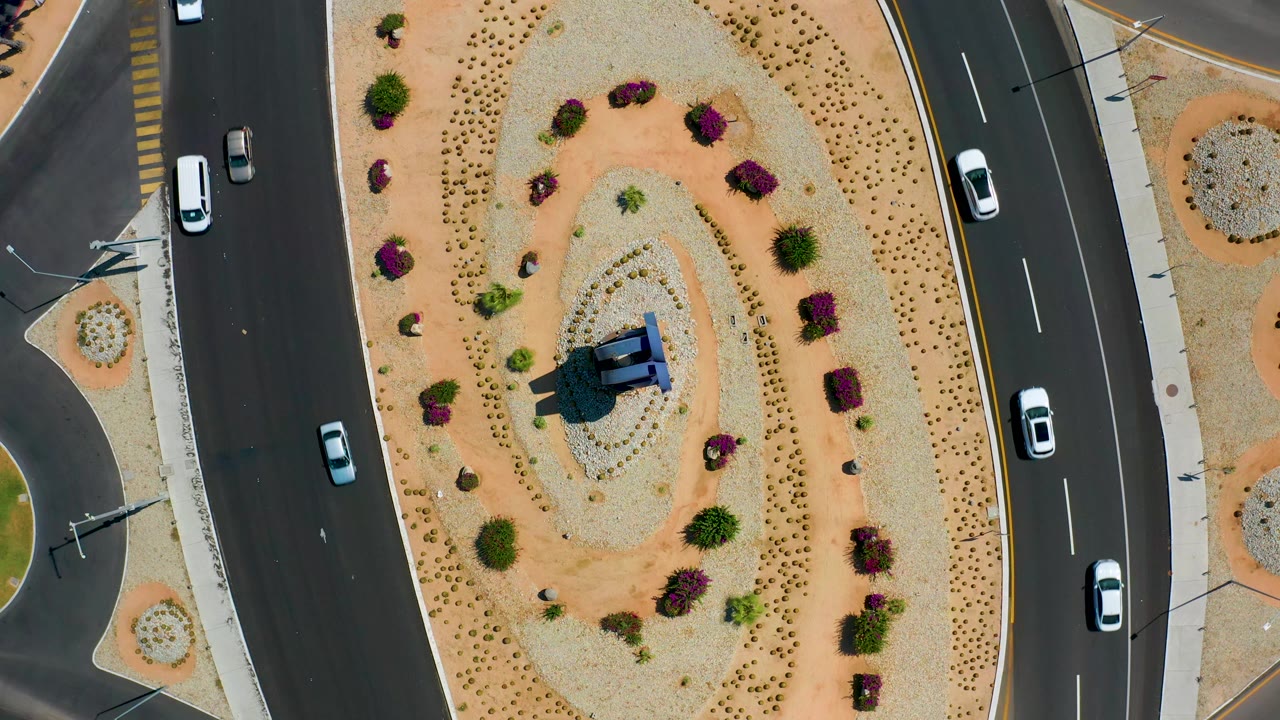 High overhead Los Cabos frame with a white car on the left road, another vehicle on the right road, and the oval planted median centered with no visible people.