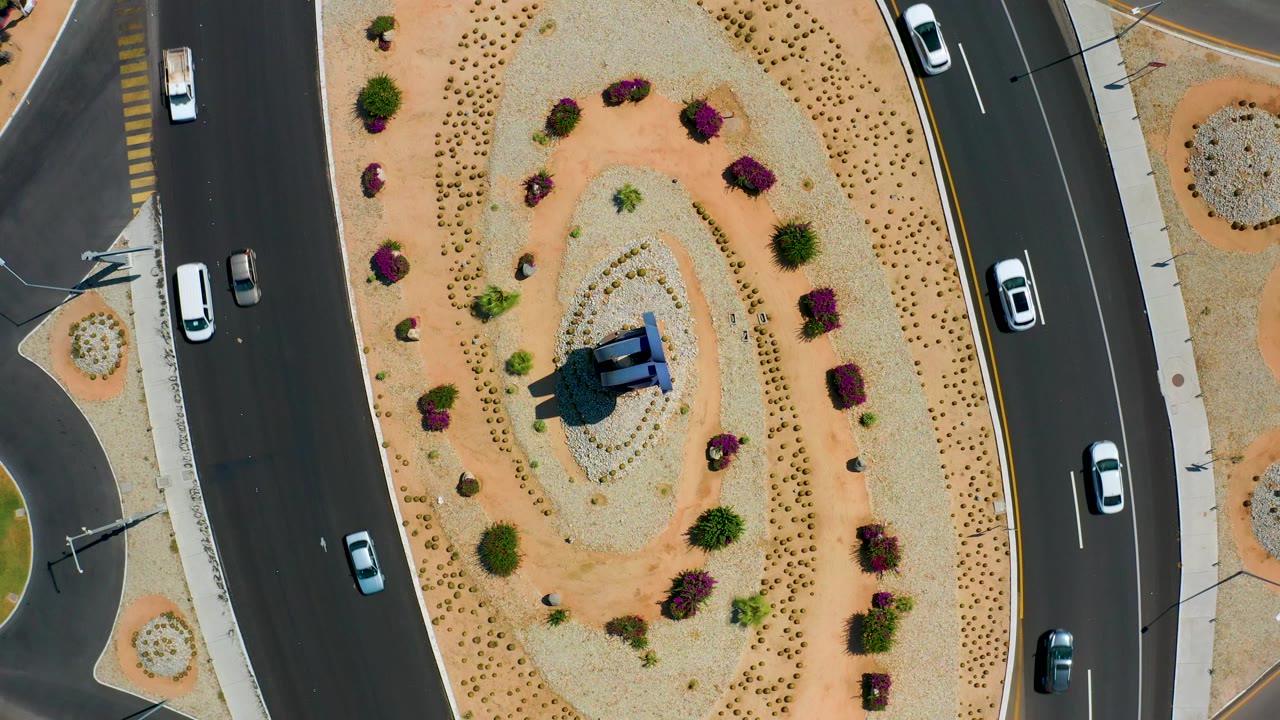 Overhead Los Cabos view of several cars on the roads around an oval desert median, with purple shrubs and pale gravel visible and no visible people.