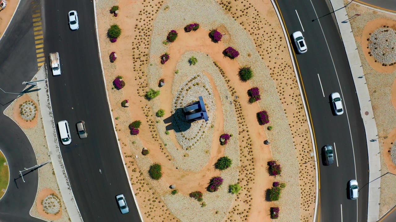 Los Cabos aerial frame looking straight down at cars on black asphalt roads bordering a planted oval median, with sidewalks and no visible people.