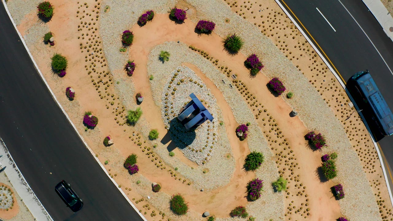 Top-down aerial frame in Los Cabos, Mexico showing a dark paved highway beside gravel and tan desert landscaping, with a bus and a car visible and no visible people.