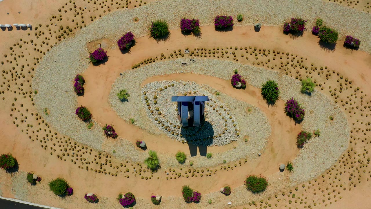 Overhead Los Cabos view with the central structure slightly lower in the oval gravel bed, curved tan sand paths around it, purple shrubs near the edges, and no visible people.