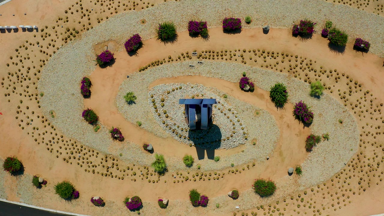 Top-down Los Cabos, Mexico frame showing a circular desert garden, pale gravel bands, purple shrubs, a central shade or sculpture-like feature, and no visible people.
