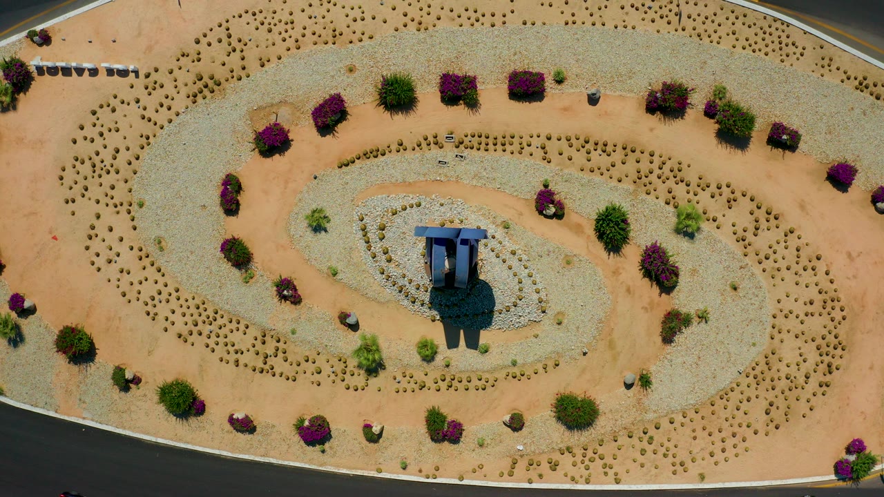 Overhead frame in Los Cabos, Mexico showing a desert landscaped oval with pale gravel, tan paths, purple shrubs, a small blue-roofed central structure, and no visible people.