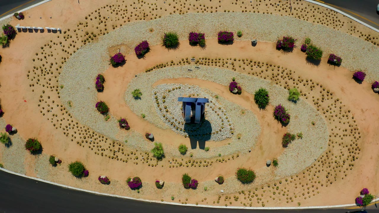 Top-down Los Cabos frame showing the landscaped island shifted slightly in view, with the blue-roofed structure near center, patterned desert plantings, roads at the edges, and no visible people.