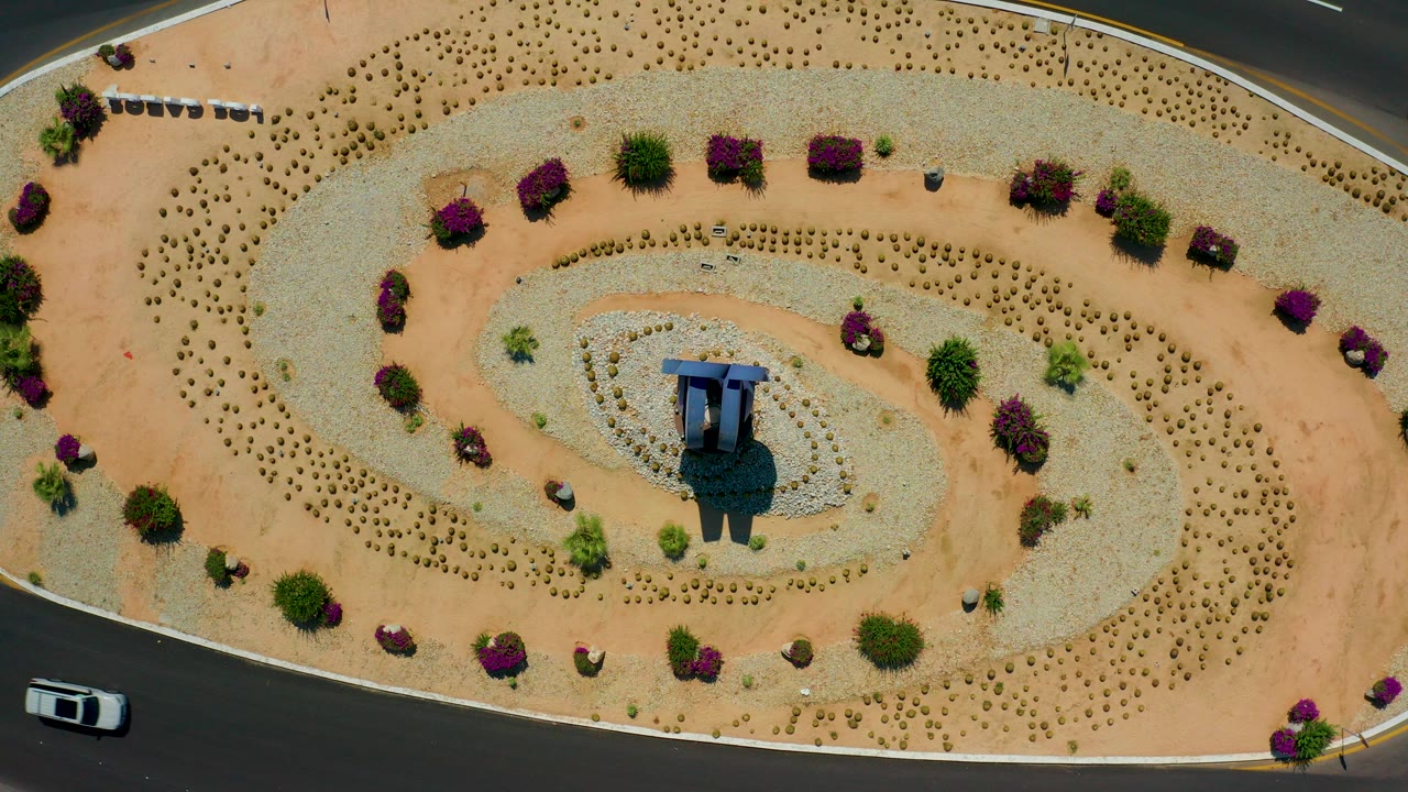 Top-down aerial frame in Los Cabos showing a spiral desert-landscaped median, a small central structure, purple shrubs, surrounding road lanes, a white car at the lower left edge, and no visible people.