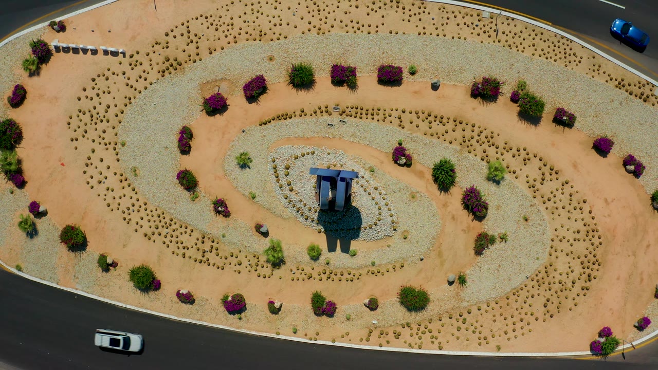 The Los Cabos overhead frame shows the same spiral median pattern with a blue car on the upper right road, a white car near the lower road, desert planting rows, and no visible people.