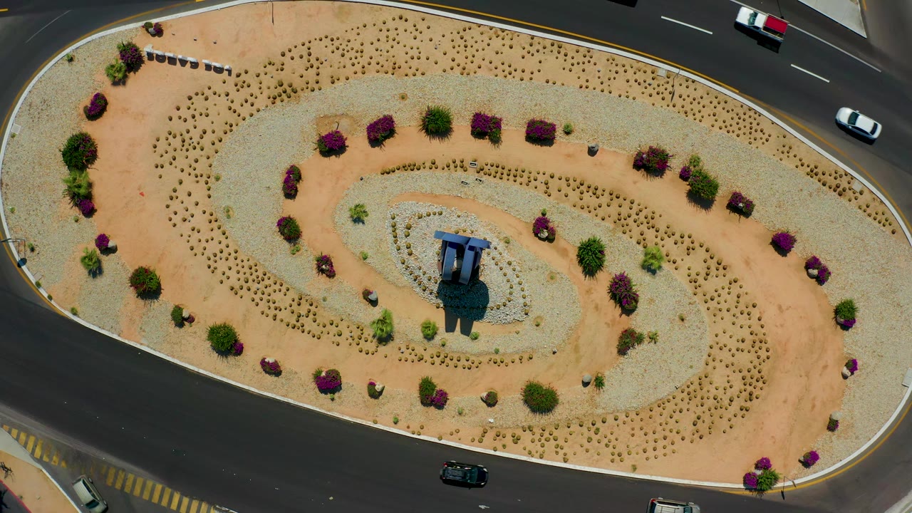 Aerial frame in Los Cabos, Mexico showing the roundabout's spiral planting pattern, purple shrubs along the curve, a blue-topped central feature, cars near the top and bottom roads, and no visible people.