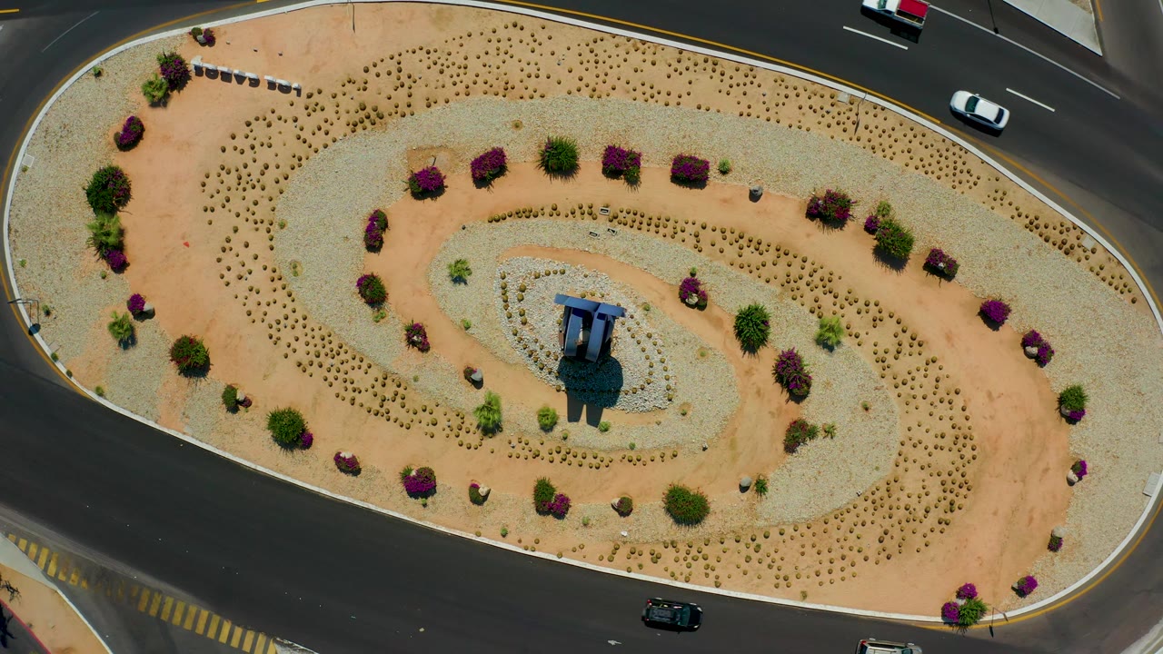 Top-down Los Cabos, Mexico view of the landscaped traffic circle with tan and pale gravel bands, scattered shrubs, a central structure, cars on the upper road, and no visible people.