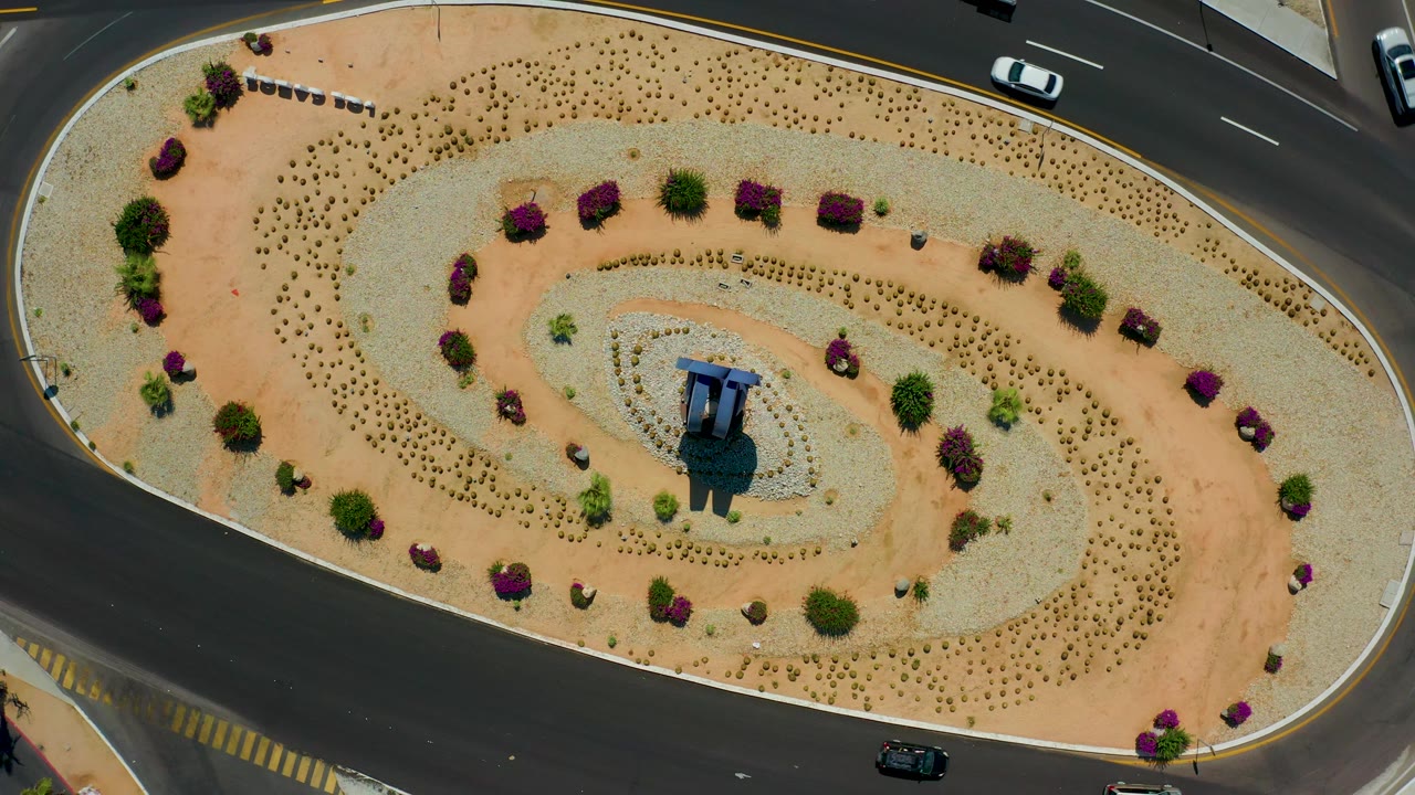Overhead Los Cabos, Mexico frame with the landscaped oval shifted slightly in view, a black car on the lower asphalt lane, a white car near the upper edge, and no visible people.