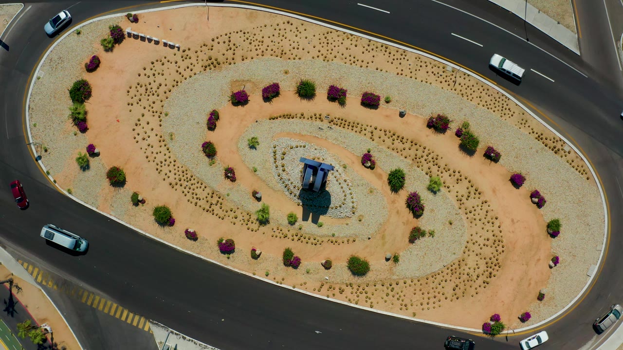 Aerial Los Cabos, Mexico frame with the roundabout still centered, a light-colored car on the upper road, several cars near the lower lanes, dry landscaping, and no visible people.