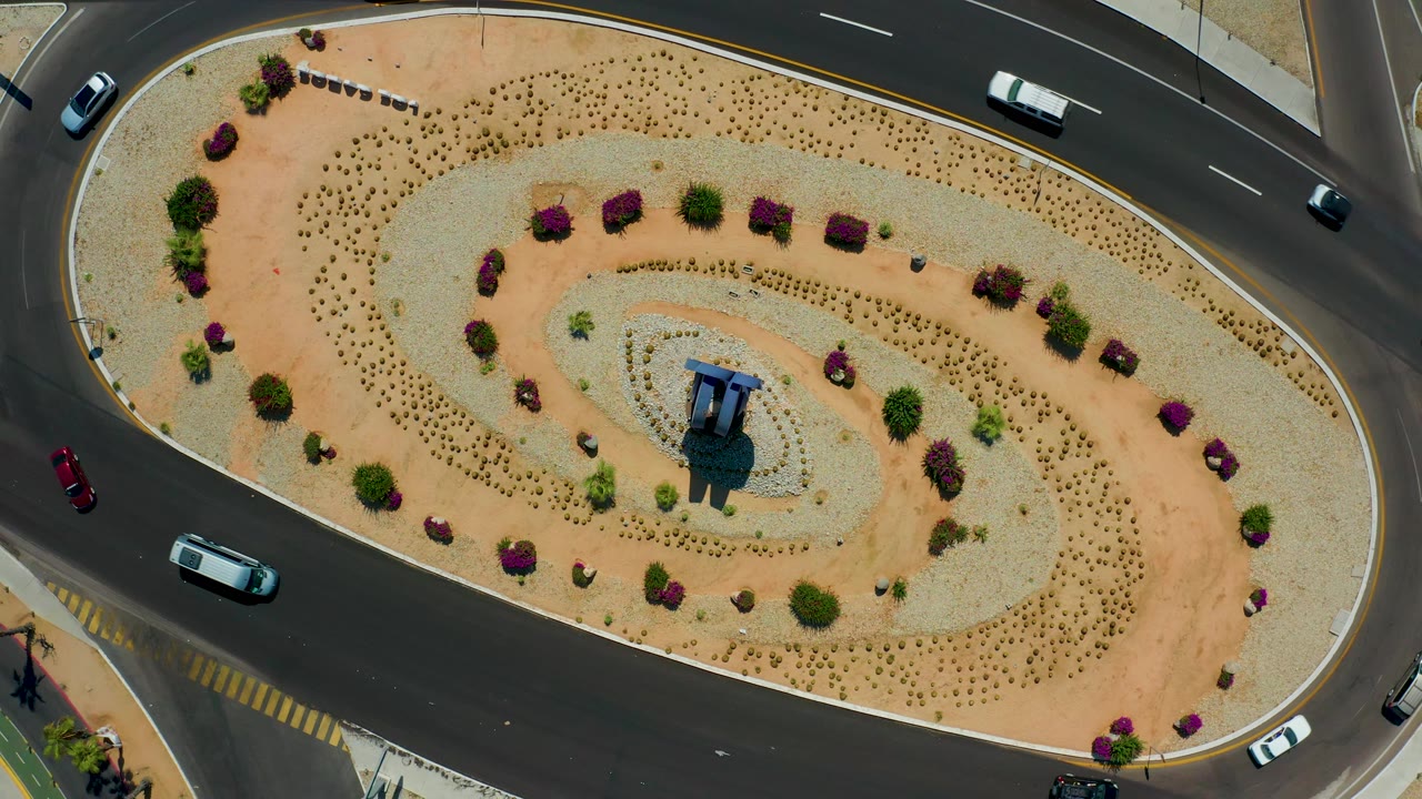 Top-down Los Cabos, Mexico frame showing cars spaced around the roundabout, a white vehicle on the upper road, the central structure casting a shadow, patterned desert planting, and no visible people.