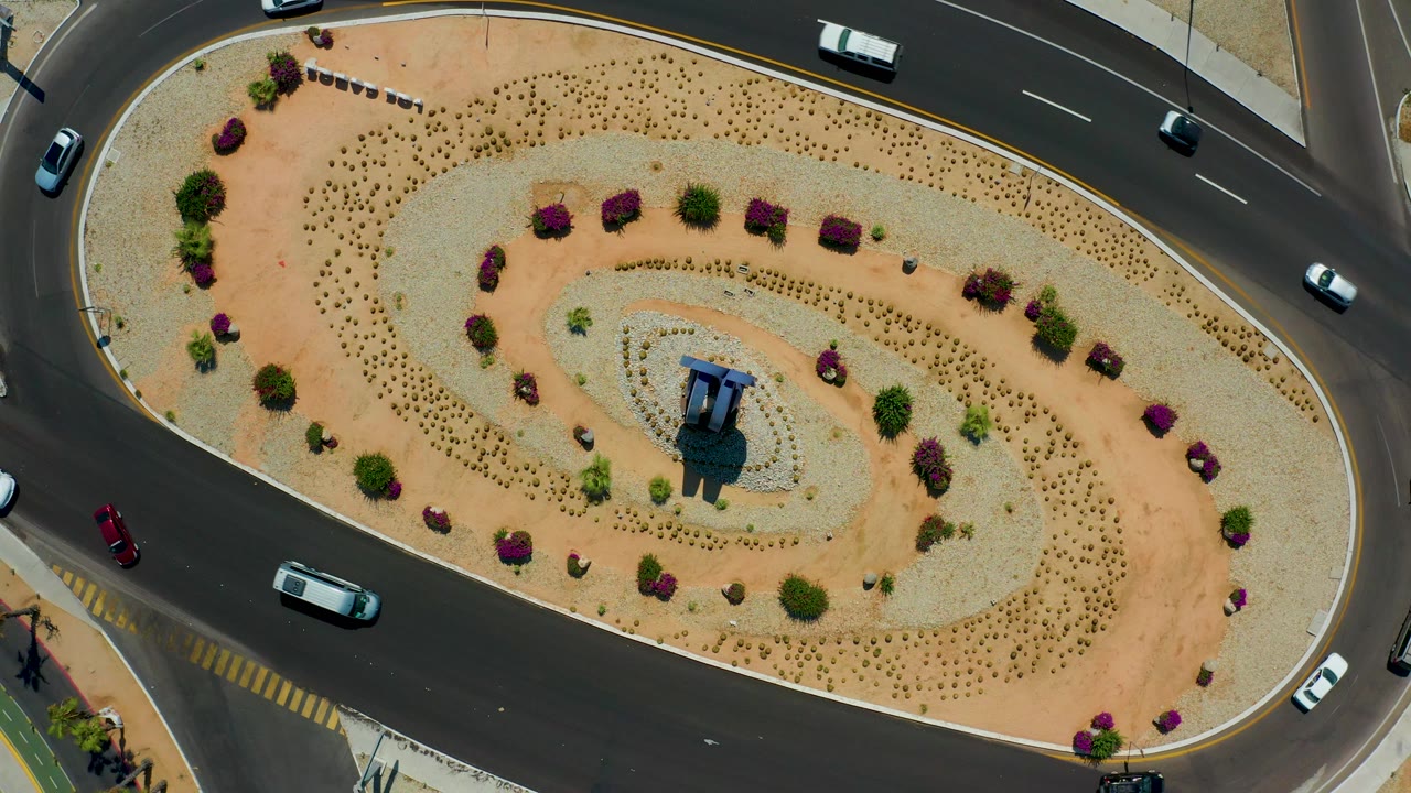 Overhead Los Cabos, Mexico frame where cars appear along the left and right road edges, the oval island holds pale gravel, sand paths, shrubs, purple plantings, and no visible people.