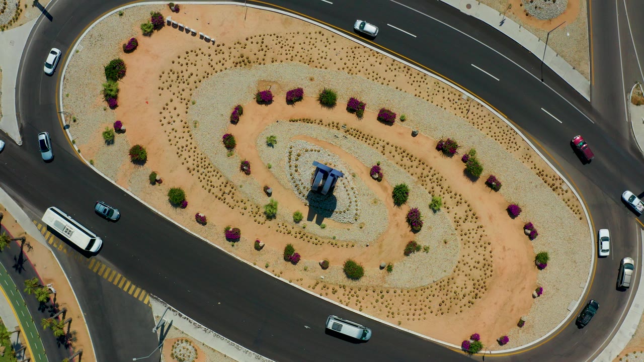 Overhead view in Los Cabos, Mexico, showing cars on curved asphalt lanes around an oval desert-landscaped island with no visible people.